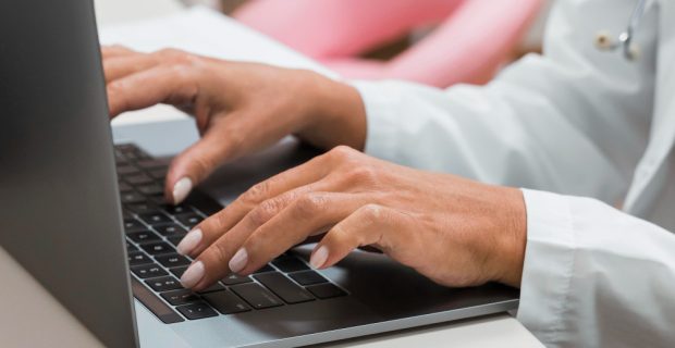 mãos de uma mulher com jaleco digitando em um notebook