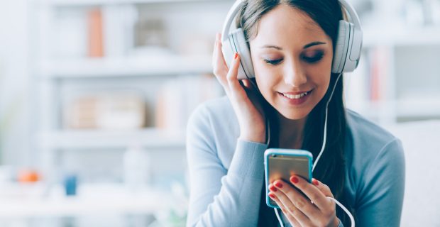 Smiling girl relaxing at home, she is playing music using a smartphone and wearing white headphones