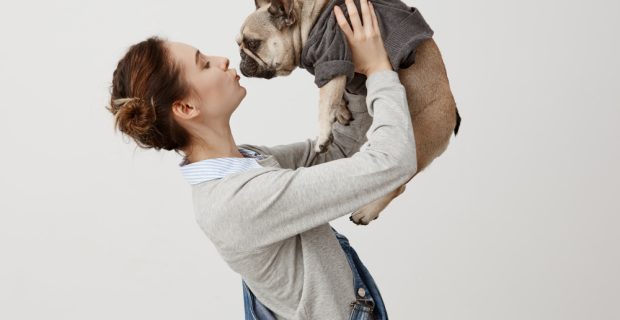 tiro-horizontal-da-menina-adulta-em-macacao-jeans-beijando-filhote-de-cachorro-bonito-enquanto-aumenta-lo-no-ar-jovem-garota-apaixonada-por-seu-cachorro-vestido-com-jumper-na-moda-demonstracao-de-carinho