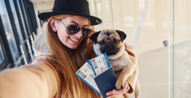 Young female passenger in warm clothes holding tickets and cute dog in hands in airport hall