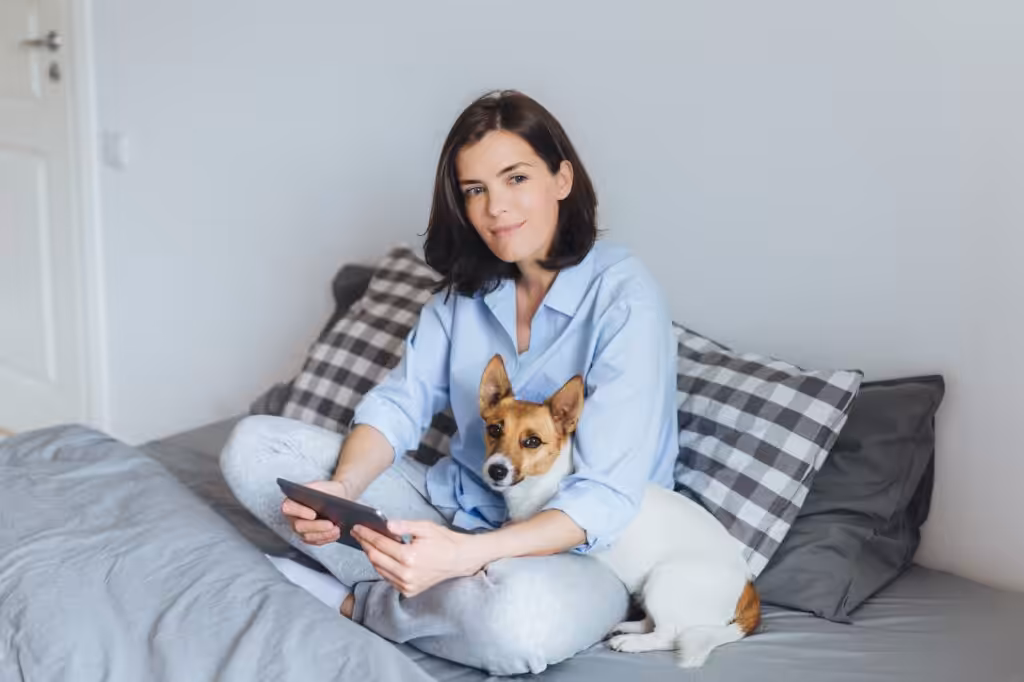 Female model in pyjamas sits on comfortable bed in bedroom, holds modern tablet, embraces her pet