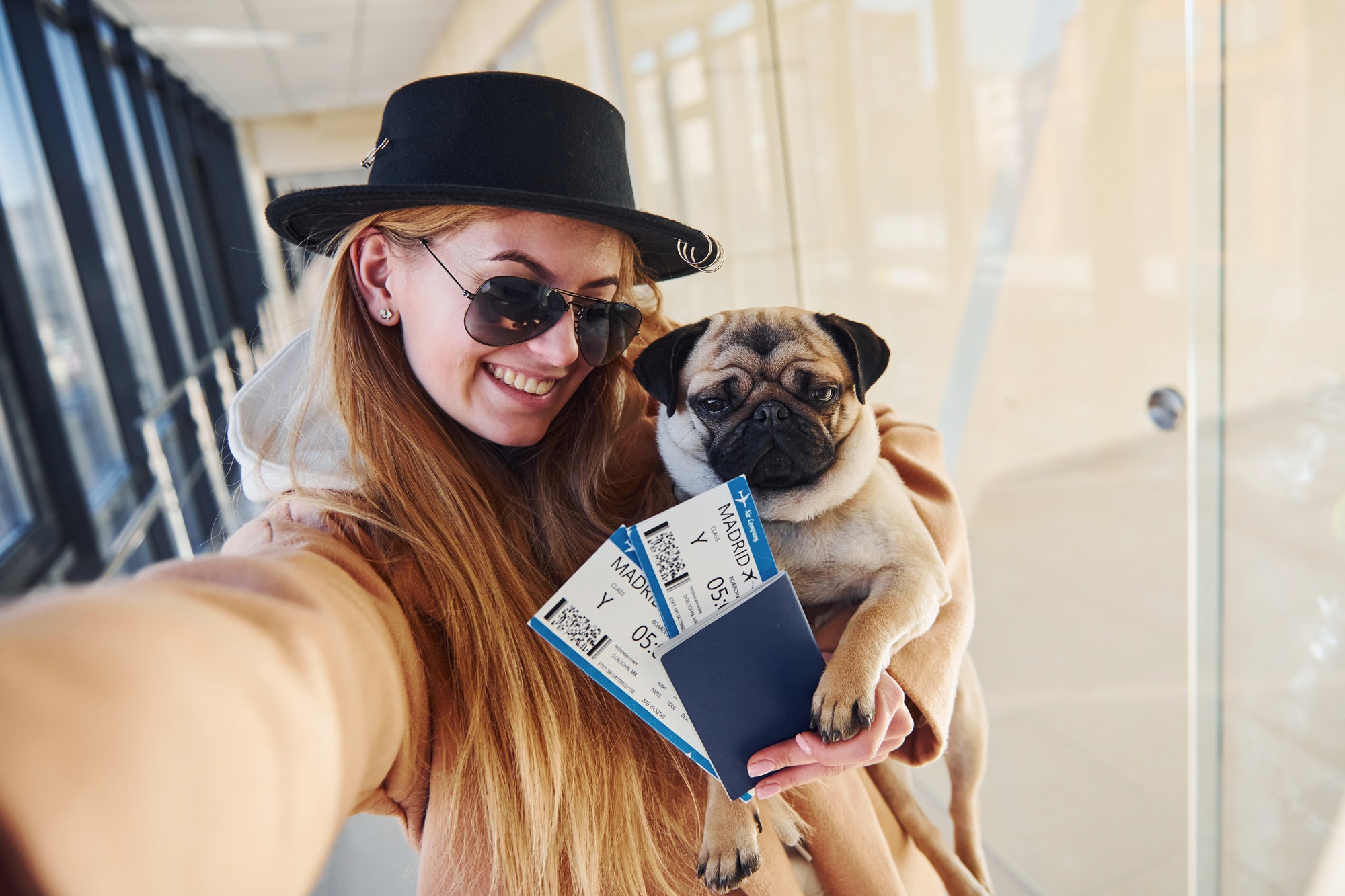 Young female passenger in warm clothes holding tickets and cute dog in hands in airport hall